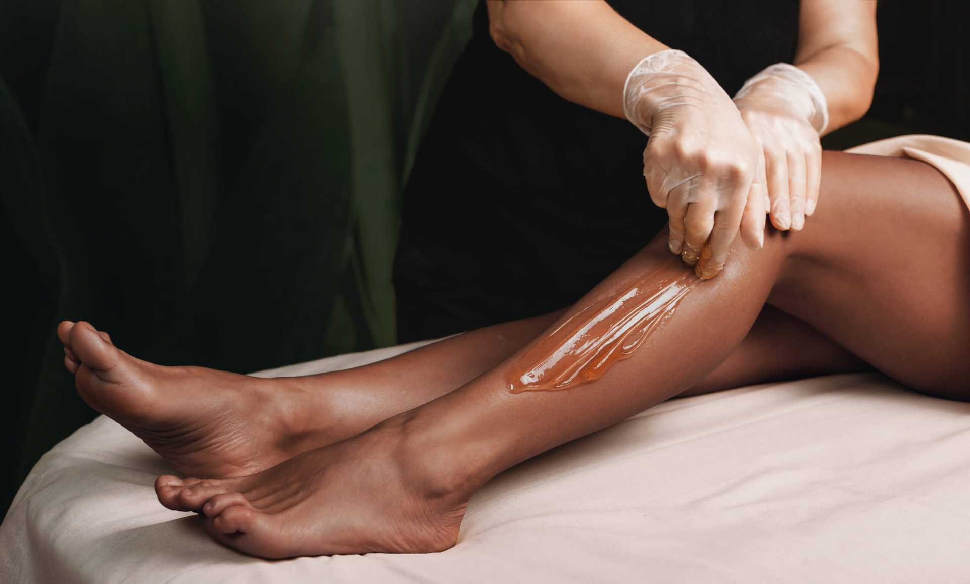 Woman applying wax to leg for hair removal on a table, wearing gloves.