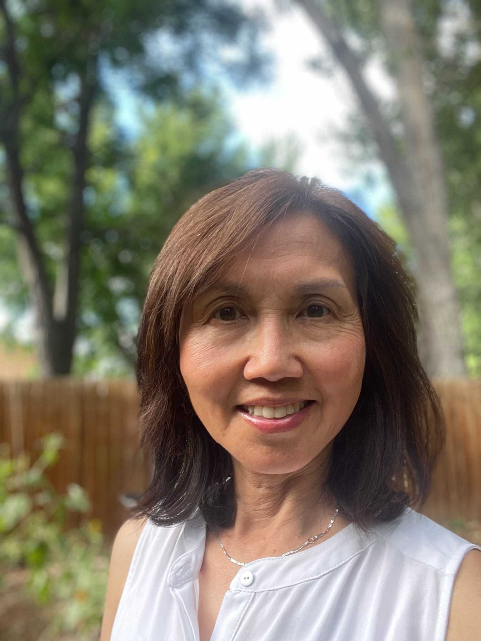 Woman smiling outdoors in a garden setting with trees and wooden fence in the background.
