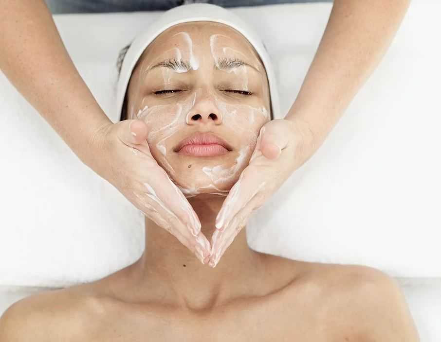 Facial treatment in progress; person relaxed with cream applied, hands massaging cheeks.