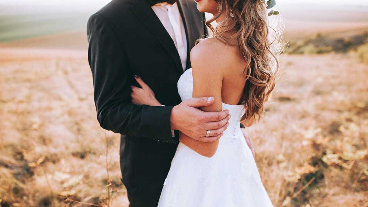 Bride and groom embracing in a field during sunset, wearing formal wedding attire.