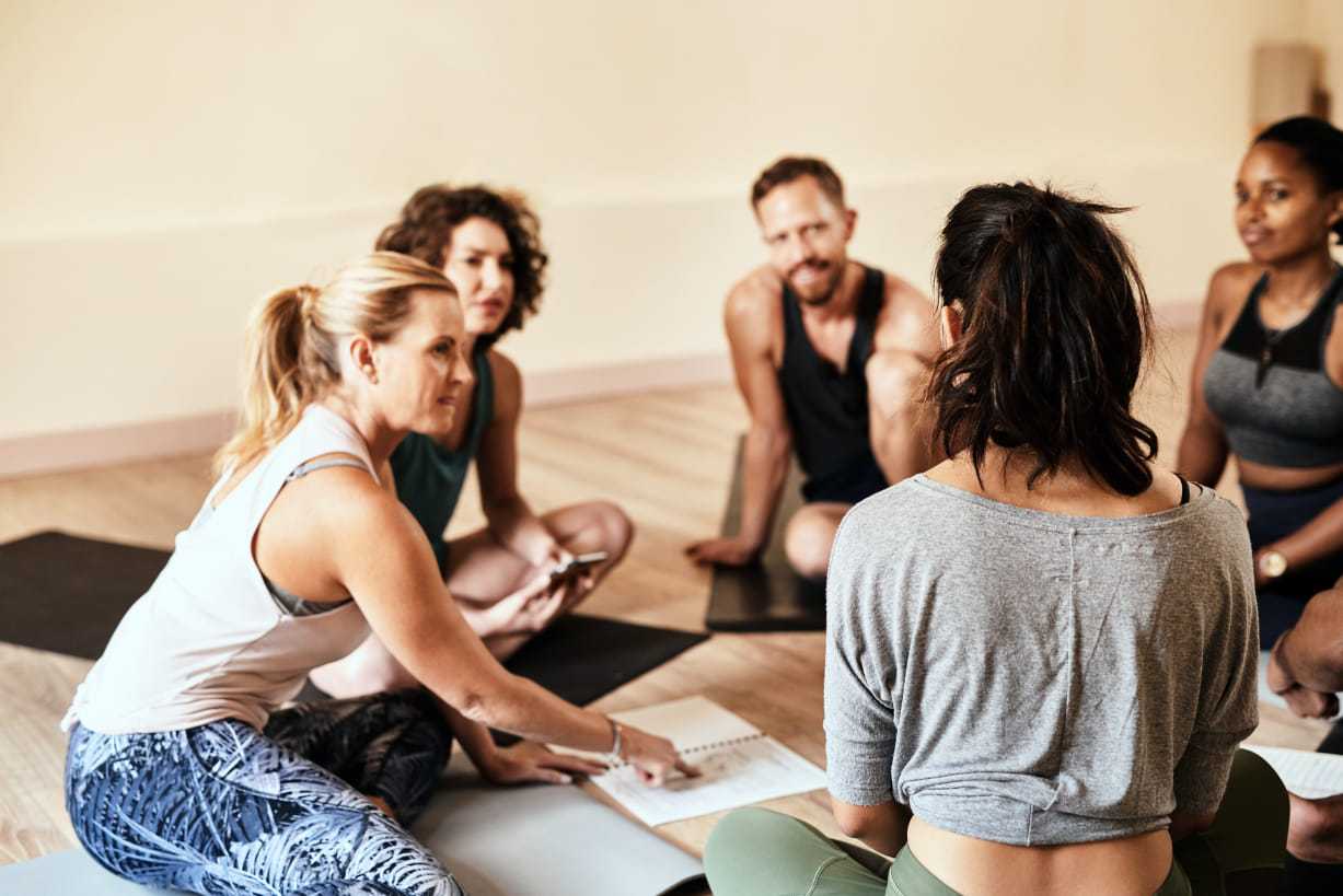 Group of people sitting on yoga mats in discussion, holding notebooks and pens.
