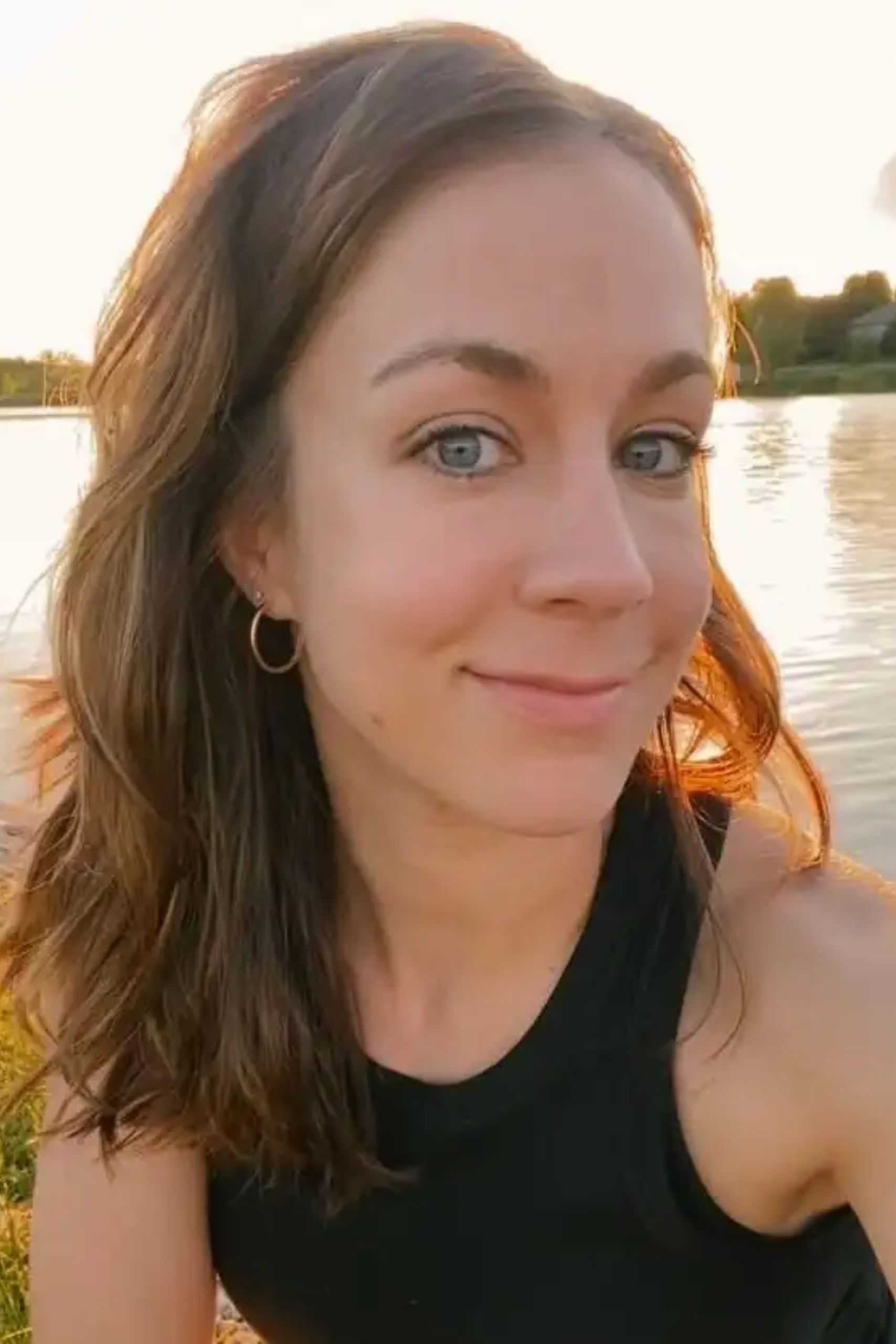 Woman smiling by a lake at sunset, with water and trees in the background.