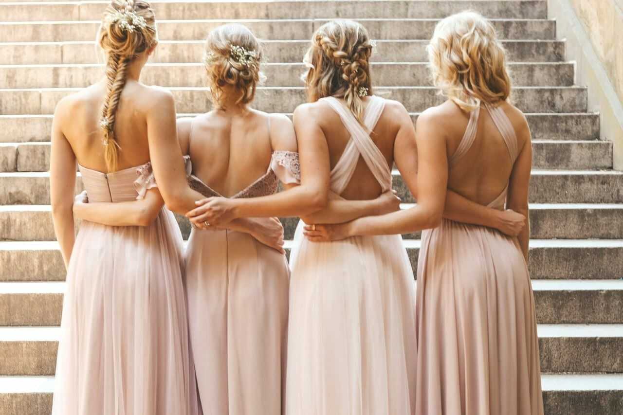 Bridesmaids in elegant pastel dresses, arms linked, standing on sunlit steps.